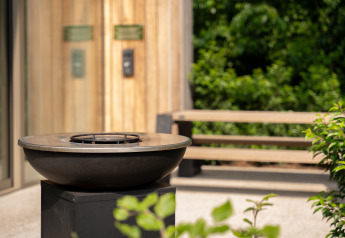 Modern outdoor fire pit at Lodge, surrounded by green foliage, bench seating, and wood-paneled wall.