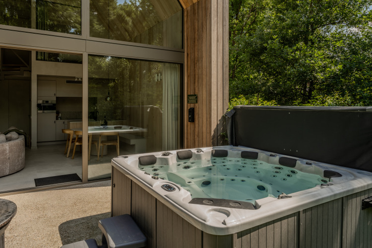 Jacuzzi al aire libre en la terraza de una moderna cabaña con grandes ventanas y vista al bosque.
