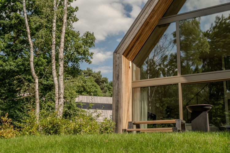 Moderna cabaña con grandes ventanales de vidrio y exterior de madera, rodeada de árboles y césped.