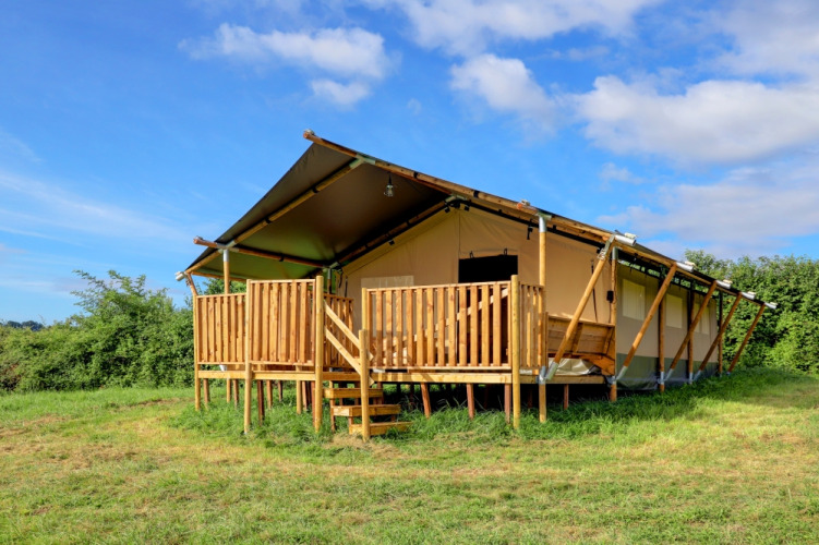 Tenda safari glamping con veranda in legno situata su un prato verde sotto un cielo azzurro, ideale per campeggi di lusso.