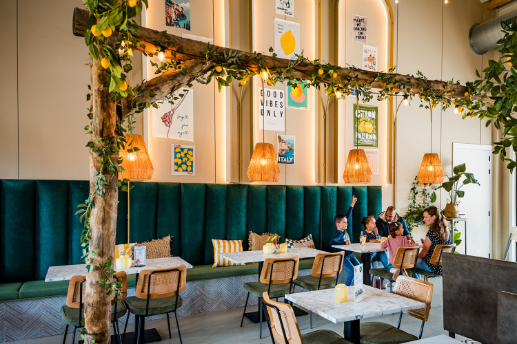 Modernes Café mit grünen Sitzbänken, Holz und Zitronendeko, Familien beim Essen zusammen.