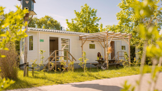 Tiny house Zomerhuisje blanche avec terrasse, végétation autour et femme assise dehors, journée ensoleillée.