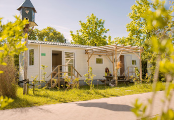 Bright Zomerhuisje tiny house with a porch, surrounded by greenery, woman sitting outside in sunlight.