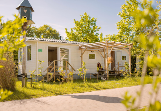 Tiny house Zomerhuisje blanche avec terrasse, végétation autour et femme assise dehors, journée ensoleillée.