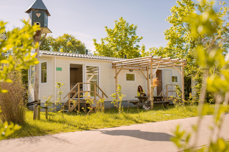 Licht zomerhuisje tiny house met veranda, omringd door groen, vrouw zit buiten in het zonlicht.
