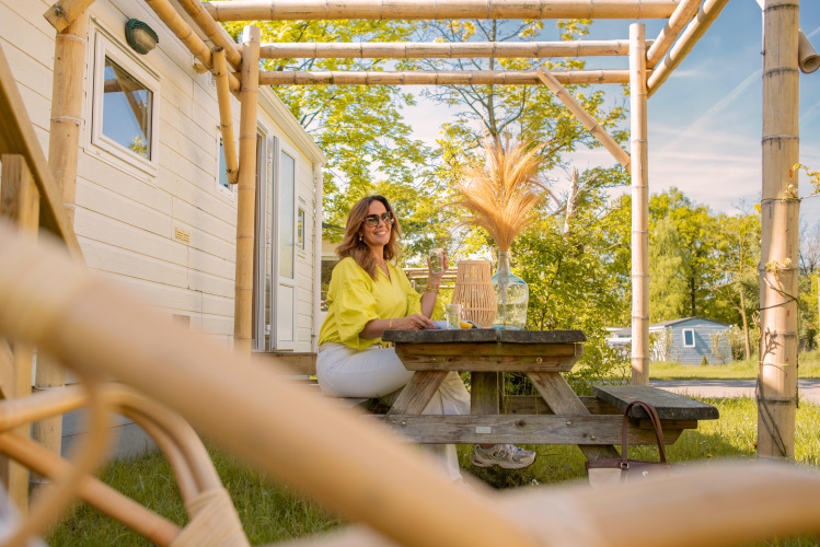 Mujer sentada en una mesa de picnic frente a una tiny house en Zomerhuisjes, Beloofde Land, Países Bajos, verano.