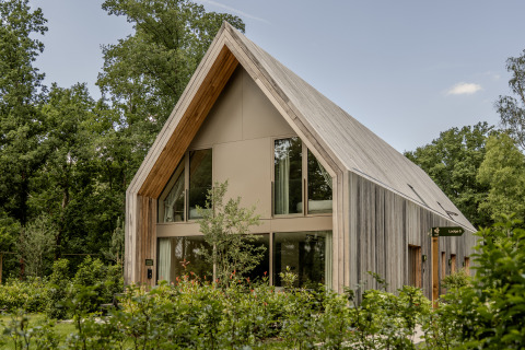 Moderne houten lodge met hoge ramen en puntdak, gelegen tussen bomen en groene beplanting.