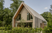 Lodge moderno in legno con grandi finestre e tetto spiovente circondato da alberi e vegetazione verde.