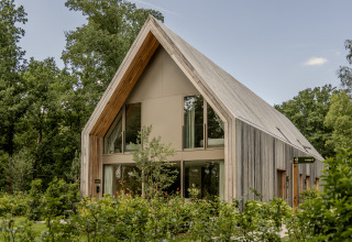 Moderne houten lodge met hoge ramen en puntdak, gelegen tussen bomen en groene beplanting.