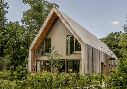 Moderne houten lodge met grote ramen en hoog puntdak, omgeven door bomen en weelderige begroeiing.
