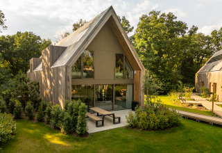 Moderne træhytte med store vinduer og terrasse, omgivet af grøn have og træer ved en lodge.