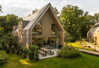 Lodge moderno con grandi finestre e patio, circondato da un giardino verde e alberi alti.