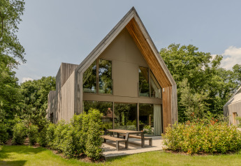 Moderne lodge med stort glasparti og træfacade, omgivet af grønne buske og træer på en solrig dag.