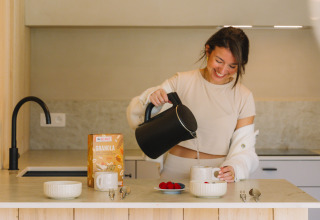 Mujer vertiendo agua de una tetera en una taza en la cocina moderna de un lodge con desayuno y granola.