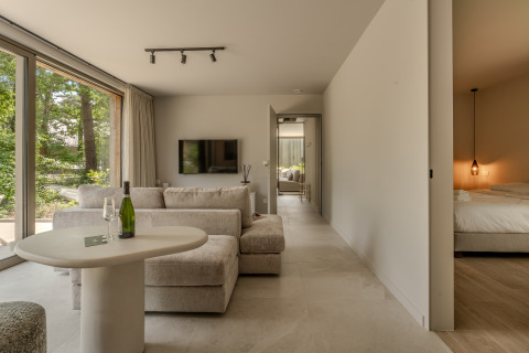 Salón moderno con grandes ventanales, sofá cómodo, mesa y vista al dormitorio en Woodz Lodges de Bélgica.