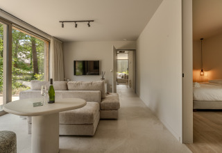 Salón moderno con grandes ventanales, sofá cómodo, mesa y vista al dormitorio en Woodz Lodges de Bélgica.