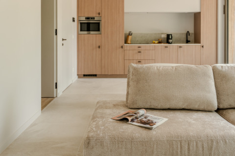 Salón moderno con sofá beige, revista y cocina visible en Group Lodge, Woodz Lodges, Bélgica.