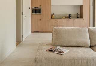 Salón moderno con sofá beige, revista y cocina visible en Group Lodge, Woodz Lodges, Bélgica.