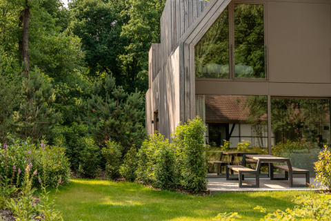 Lodge moderno de madera junto a un jardín verde y frondoso con bancos y mesas al aire libre en el bosque.