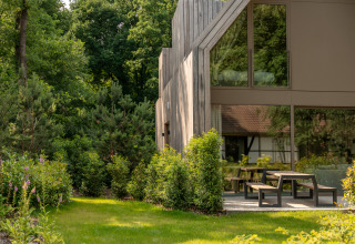 Lodge moderno de madera junto a un jardín verde y frondoso con bancos y mesas al aire libre en el bosque.