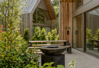 Lodge moderne avec bardage en bois, grandes fenêtres, verdure abondante et espace détente en plein air.