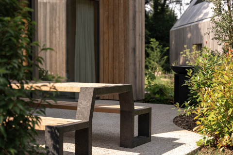 Banco y mesa de exterior en una casa de campo con fachada de madera, rodeados de plantas verdes.