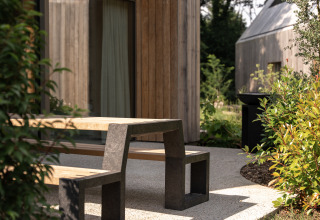 Banco y mesa de exterior en una casa de campo con fachada de madera, rodeados de plantas verdes.