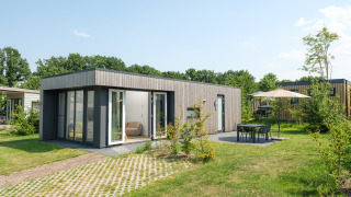 Moderne Oak Lodge mit großen Fenstern, Terrasse, Gartenmöbeln und Sonnenschirm an einem sonnigen Tag.