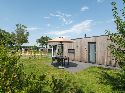 Moderne træhytte med terrasse, havemøbler og parasol på Vakantiepark Hölte i Holland under blå himmel.