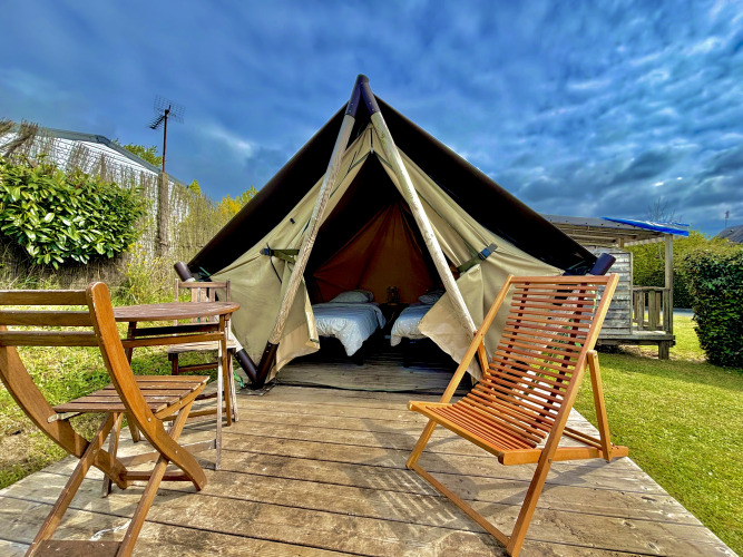 Nomad Bivouac Tipi-Zelt mit hölzernem Deck, Stühlen und Tisch auf einer grünen Wiese bei bewölktem Himmel.