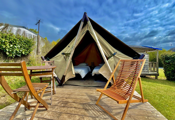 Tienda tipi Nomad Bivouac con terraza de madera, sillas y mesa en un jardín verde bajo un cielo nublado.