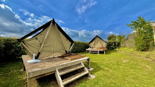 To glamping-telte på forhøjede trækonstruktioner i en grøn have under en blå himmel med skyer.