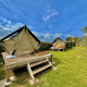To glamping-telte på forhøjede trækonstruktioner i en grøn have under en blå himmel med skyer.