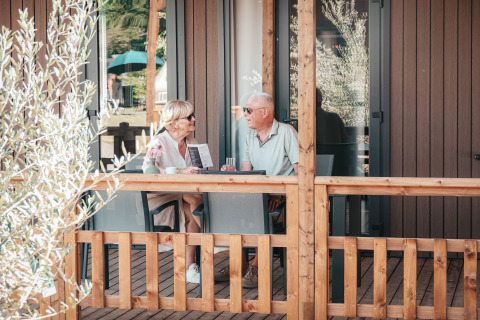 Couple senior assis et discutant sur la terrasse en bois du Tiny Wander Lodge lors d'une journée ensoleillée.