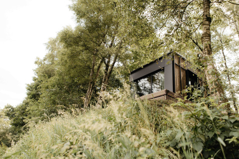 The Nokken NKN-18 Cabin, a modern tiny house, nestled on a hillside among trees and lush greenery in nature.
