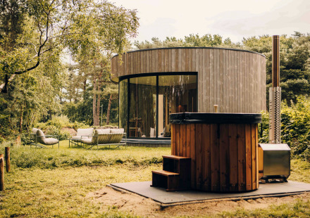 Ronde LumiPod tiny house met grote ramen, houten gevel en hottub, omgeven door groen landschap.