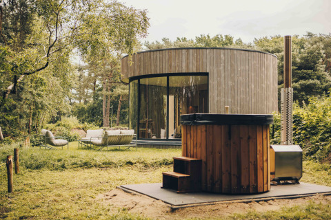 Ronde LumiPod tiny house met grote ramen, houten gevel en hottub, omgeven door groen landschap.
