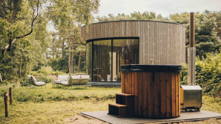 Ronde LumiPod tiny house met grote glaspartijen, houten bekleding en hottub in een bosrijke omgeving.