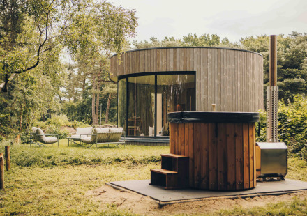 Ronde LumiPod tiny house met grote ramen, houten gevel en hottub, omgeven door groen landschap.