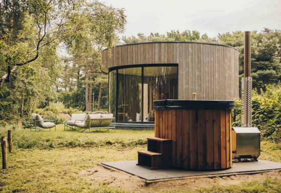 Ronde LumiPod tiny house met grote ramen, houten gevel en hottub, omgeven door groen landschap.