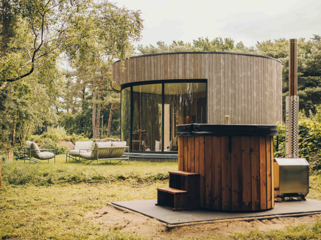 Ronde LumiPod tiny house met grote ramen, houten gevel en hottub, omgeven door groen landschap.