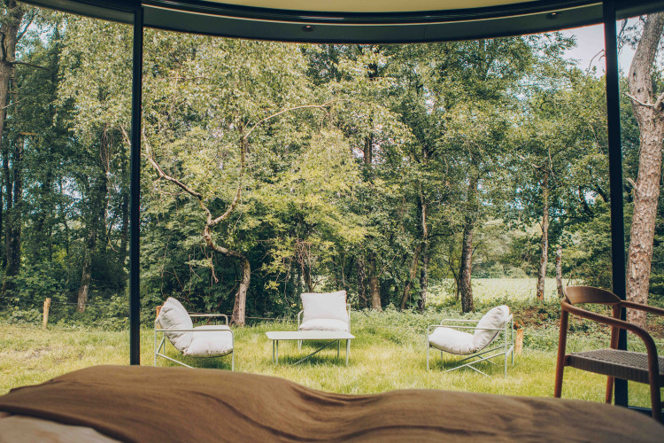 View from inside a tiny house with large windows to outdoor chairs, a table, and forest at Wilsumer Berge, Germany.