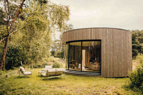 Rundes, holzverkleidetes Tiny House mit großer Fensterfront und Gartenmöbeln in Wilsumer Berge, Deutschland.
