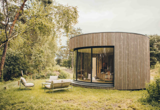Rond, houten tiny house met grote glazen pui en buitenzitjes in het groen bij Wilsumer Berge, Duitsland.