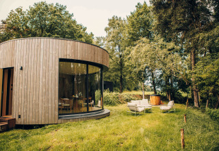 Rundes Tiny House aus Holz mit großer Fensterfront, gemütlichen Sitzmöbeln und Hot Tub im grünen Waldgebiet.
