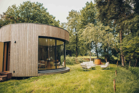 Rond houten tiny house met grote ramen, comfortabele tuinset en een hottub in een groene bosrijke omgeving.