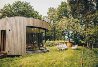 Rond houten tiny house met grote ramen, comfortabele tuinset en een hottub in een groene bosrijke omgeving.