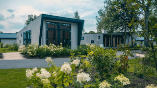 Lodge moderne Just Nature 4 avec de grandes fenêtres, entourée de verdure et de fleurs blanches en fleurs.