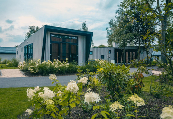 Lodge moderne Just Nature 4 avec de grandes fenêtres, entourée de verdure et de fleurs blanches en fleurs.