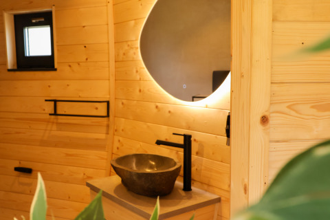 Modern badkamer in Wellnest, Camping Vinkenhof, Nederland, met houten wanden, ronde spiegel en wastafel.